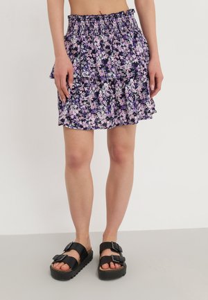 Jupe fleurie avec une base violet foncé et des fleurs pastel, composée d'un ourlet à volants et d'une taille élastique. Associée à des sandales noires à double boucle.