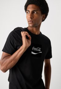 Black cotton t-shirt featuring a white Coca-Cola logo graphic on the front. Short sleeves and standard crew neck design.