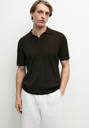 Giovane uomo con capelli chiari che indossa una polo a maniche corte in maglia marrone scuro e pantaloni bianchi, in piedi davanti a uno sfondo bianco semplice.