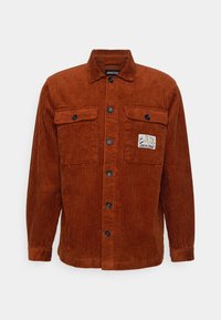 Rostfärgad corduroy skjortjacka med krage, två bröstfickor och knäppning. Har en märke på den vänstra fickan.