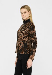 Giacca con stampa leopardata in marrone e nero, presenta un alto colletto, chiusura a zip e un tessuto morbido con una texture liscia. Accentuata con accessori in oro.
