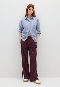 Camicia a righe blu e bianche con bottoni, abbinata a pantaloni larghi bordeaux con strisce laterali bianche. Il modello si trova di fronte a uno sfondo neutro.