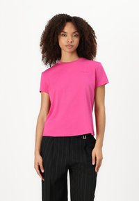 T-shirt a girocollo in cotone rosa con maniche corte, caratterizzato da un sottile logo del marchio sul petto. Indossato con pantaloni neri a righe sottili.