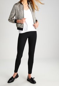 Donna con una giacca bomber argentata, top bianco, pantaloni neri slim e mocassini neri con frange, in piedi davanti a uno sfondo semplice chiaro.