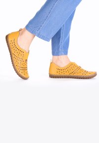 Gelbe Leder Slipper mit Ausschnittmustern, strukturierten Kanten und einer braunen Gummisohle, kombiniert mit hellblauen Jeans.