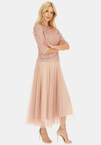 Różowa złota bluzka z cekinami i rękawami o długości trzech ćwiartek, zestawiona z zwiewną, przezroczystą spódnicą z jasnoróżowego tiulu oraz cielistymi, sandałowymi szpilkami.