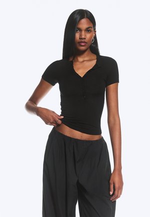 Kvinde med glat sort hår iført en tætsiddende sort kortærmet bluse med knapper og løse sorte bukser, som løfter sin bluse let med den ene hånd.