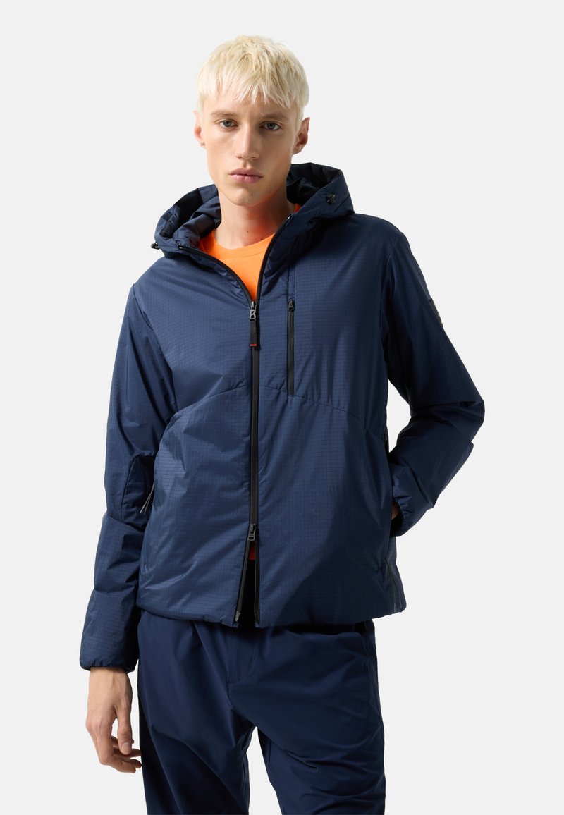 Young person wearing a navy blue hooded jacket over an orange shirt, with navy blue pants, standing against a light gray background.