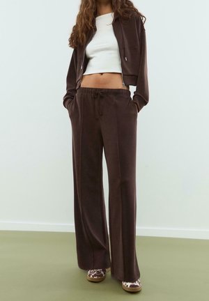 Nainen, jolla on tummanruskeat leveälahkeiset housut kiristysnauhalla, yhteensopiva lyhyt vetoketjullinen takki, valkoinen lyhyt toppi ja lehmäkuvioidut kengät.