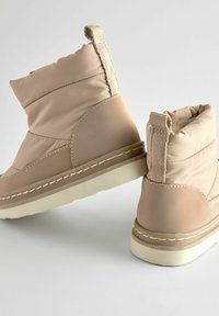Lichtbeige gevoerde laarzen met een gladde afwerking, voorzien van een getextureerde trekband en witte stiksels op een crème kleurige zool.