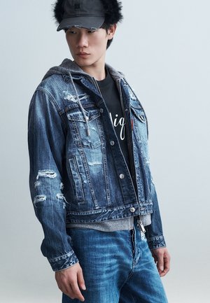 Νεαρός άνδρας με μαύρο καπέλο, γκρι φούτερ με στρώσεις, φθαρμένο μπουφάν από τζιν, μαύρο μπλουζάκι με στάμπα και μπλε τζιν, να στέκεται μπροστά σε απλό φόντο.