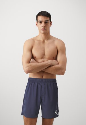 Marineblaue Badeshorts mit elastischem Bund, ausgestattet mit einer Seitentasche und weißen Logo-Details. Der Stoff wirkt leicht und glatt. Benutzer mit verschränkten Armen.