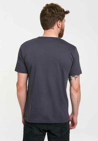 Man with short brown hair and beard wearing a plain dark gray T-shirt and black jeans, facing away showing tattoos on left arm.