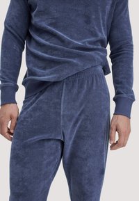 Nahaufnahme einer Person in einem passenden, weichen blauen Frottee-Oberteil mit langen Ärmeln und elastischer Bundhose, die Hände entspannt an den Seiten.