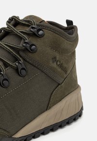 Acercamiento a una bota de senderismo Columbia de color verde olivo con ganchos metálicos negros para los cordones, suela texturizada y paneles de ante y malla.