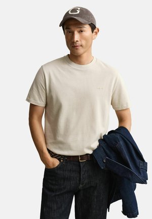 Man wearing beige t-shirt and grey cap, holding a blue denim jacket over one arm, with hand in pocket, standing against white background.
