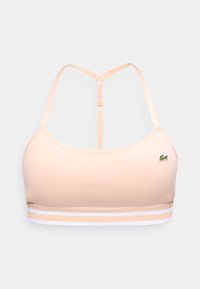 Beige ribbed sports bra with white striped elastic band and adjustable T-back straps, featuring small green crocodile logo on front.