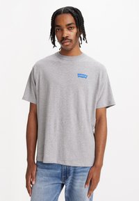 Camiseta de algodón gris de manga corta, con cuello redondo y un parche del logo azul de Levi's en el lado izquierdo del pecho. Lleva puestas unos vaqueros de color azul claro.