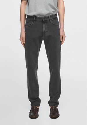 Person trägt dunkelgraue, gerade geschnittene Jeans, graues, in die Hose gestecktes T-Shirt und dunkelbraune Lederschlupfschuhe vor einfarbigem Hintergrund.