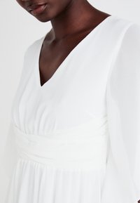 Λευκό φόρεμα με V-λαιμόκοψη, συγκεντρωμένο μπούστο και μακριά, διάφανα μανίκια. Το υλικό φαίνεται ελαφρύ και ρο flowing με λεία υφή.