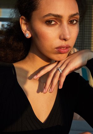 Srebrne kolczyki w kształcie prostokąta z przezroczystymi kamieniami. Na dłoni znajduje się pierścionek z zakrzywioną obrączką oraz centralnym przezroczystym kamieniem.