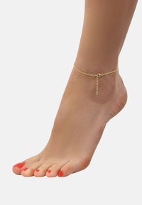 Tobillera de cadena de oro con un diseño de eslabones finos, que presenta un cierre ajustable y un pequeño acento de cadena colgante. El pie tiene esmalte rojo en las uñas.