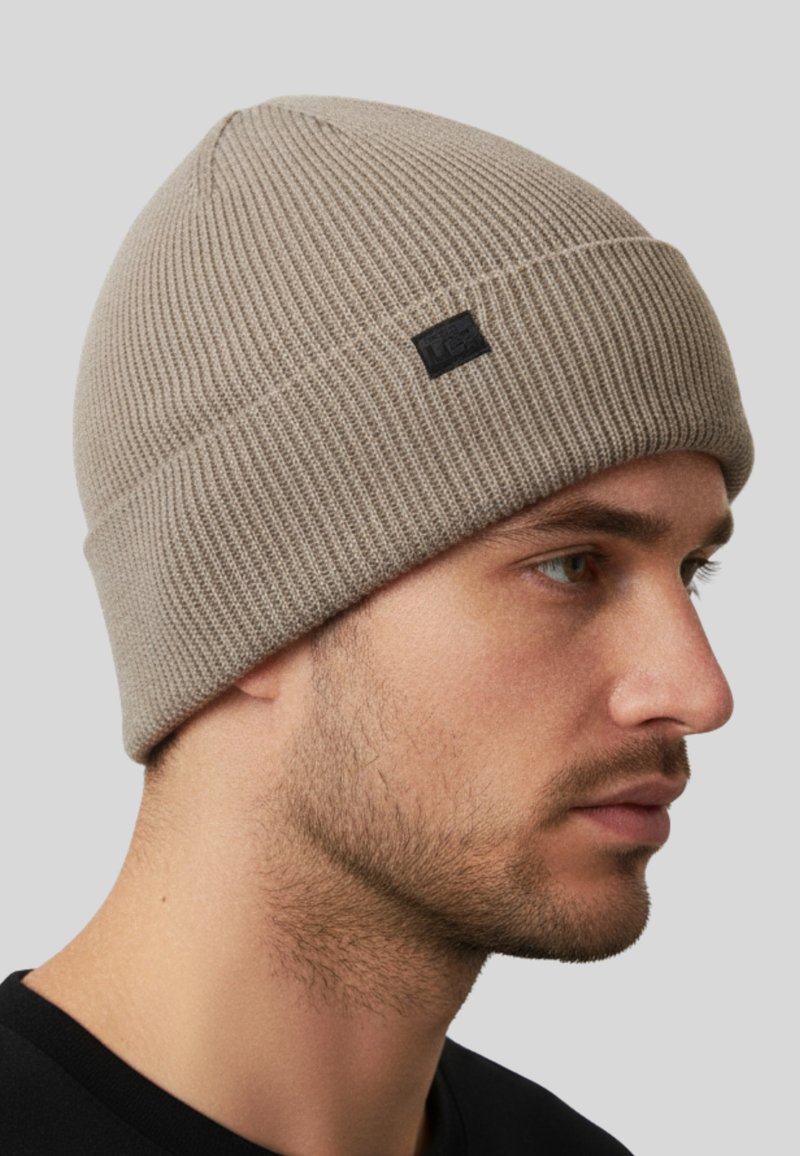 Uomo che indossa un berretto a costine beige con una piccola etichetta nera, rivolto verso destra, su uno sfondo grigio uniforme.