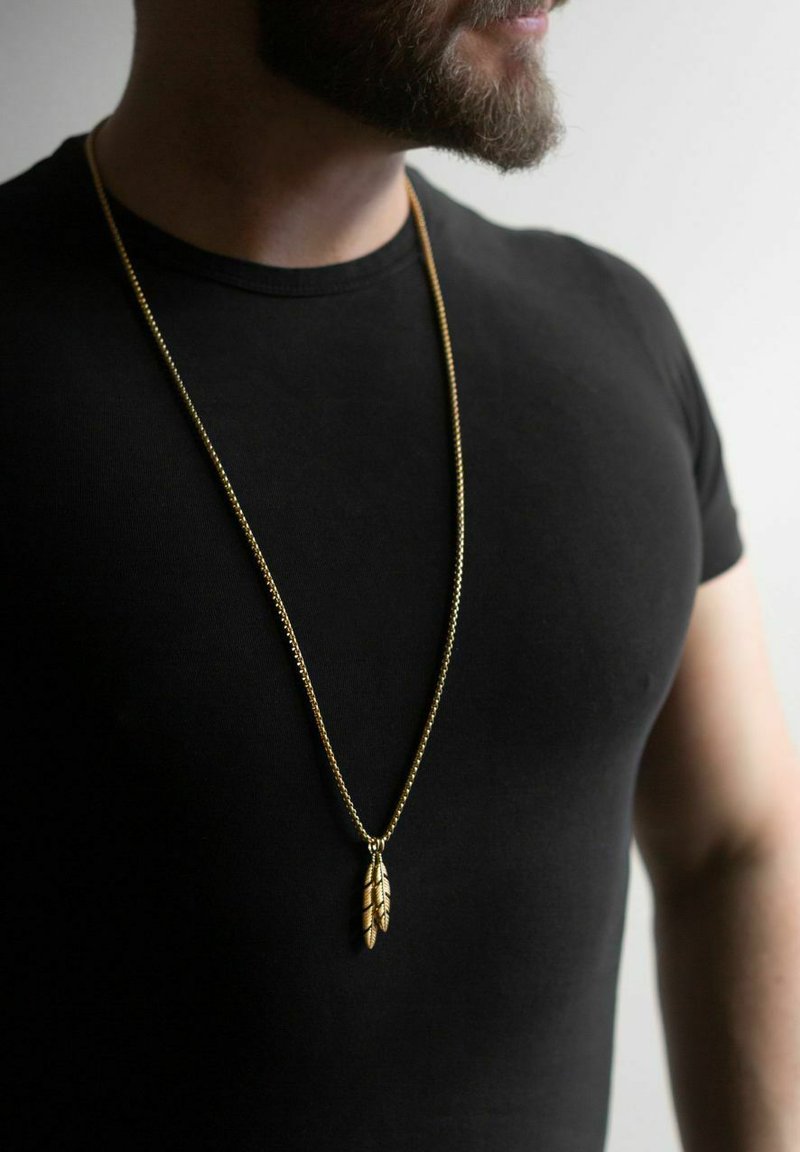 Goldene Kettenhalskette mit einem Anhänger in Form von Federn, getragen über einem schwarzen Hemd. Die Kette hat ein dünnes, strukturiertes Design.