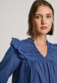 Blusa blu in tessuto texturizzato con dettagli arricciati e scollatura a V. Caratterizzata da dettagli plissettati lungo il petto e maniche lunghe.