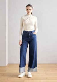 Cămașă bej cu mâneci lungi și un design răsucit, asociată cu blugi largi de culoare albastru închis, cu margini brut și mansete din denim albastru deschis. Adidași albi.