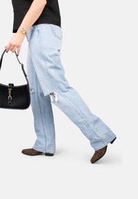 Jeans déchirés bleu clair avec une coupe décontractée, présentant des déchirures aux genoux. Associés à un sac à main noir et des chaussures à talons pointus en imprimé léopard.
