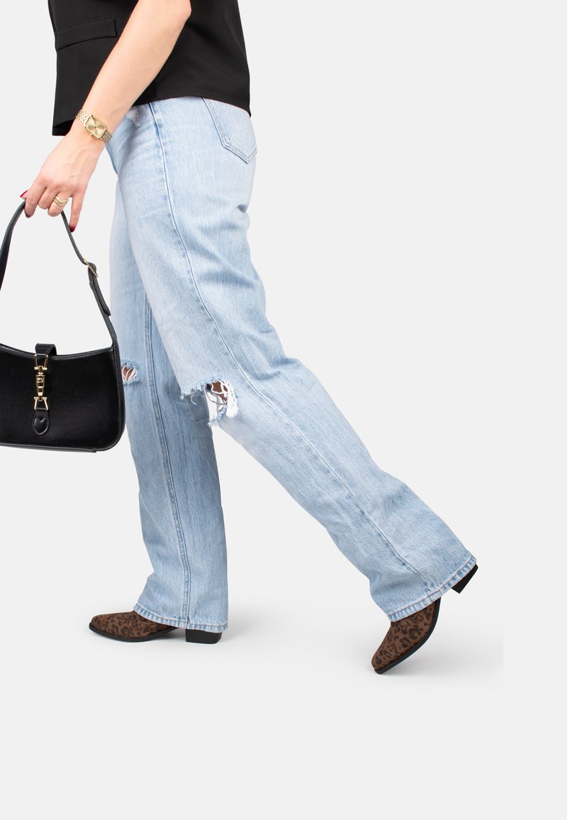 Jeans déchirés bleu clair avec une coupe décontractée, présentant des déchirures aux genoux. Associés à un sac à main noir et des chaussures à talons pointus en imprimé léopard.