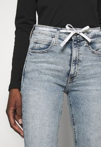 Ljusblå denimjeans med en tight design, som har en vit märkesmidja och fickor. Subtil fading ger textur.