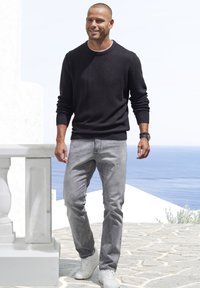 Schwarzer Strickpullover, graue Jeans und weiße Sneakers, getragen von einem Mann. Steht auf einem steinernen Untergrund mit einem Meer im Hintergrund und wirkt lässig.