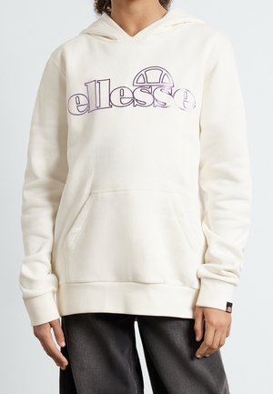 Person iført en off-white hættetrøje med en stor lilla "ellesse"-logo og frontlomme, kombineret med mørke bukser.