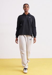 Fekete, hosszú ujjú póló, gallérral, világosszürke melegítőnadrággal párosítva, oldalcsíkkal és logóval. A modellen fehér sportcipő van.