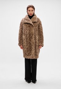 Καφέ φ furry παλτό με φαρδύ γιακά, κλείσιμο με κουμπί και υφασμάτινη υφή, συνδυασμένο με μαύρα παντελόνια και μυτερές γόβες.