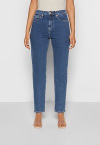 High-waisted, straight-leg blue denim jeans with subtle fading, five pockets, and stitching accents, paired with clear slide sandals.