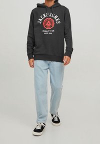 Schwarzer Hoodie mit weißem und rotem grafischen Text, kombiniert mit hellblauen Jeans und schwarzen Sneakers. Der Hoodie hat einen Kordelzug und eine Vorderseite mit Tasche.