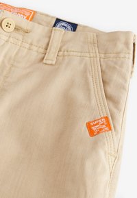 Pantaloni beige realizzati in un tessuto testurizzato con cuciture visibili, dotati di un'etichetta "Superdry" arancione sul davanti e chiusura a bottone.