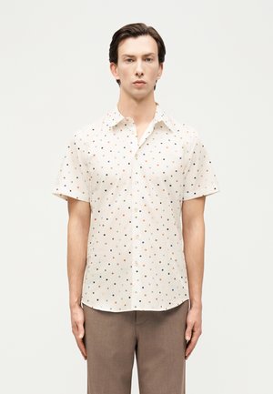 Άνδρας που φοράει κοντομάνικο λευκό πουκάμισο με μικρό πολύχρωμο μοτίβο τετραγώνων και καφέ παντελόνι, στέκεται με τα χέρια στα πλευρά.