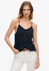 Navy blauwe peplum top met dunne bandjes, een V-hals, haakwerk accenten en een gestructureerde stof. Gestyled met een gevlochten riem en lichte broek.