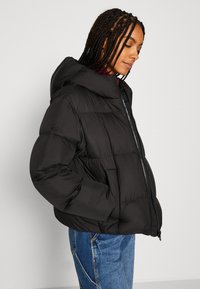 Schwarze Daunenjacke mit Kapuze, ausgestattet mit einem gesteppten Design, Vorderreißverschluss und seitlichen Taschen. Leicht taillierter Schnitt.