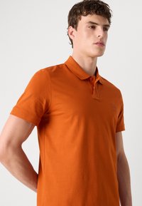 Kortermet polo-skjorte i livlig oransje, laget av glatt bomullsstoff. Har en klassisk krage og diskret logo-detaljering på brystet.