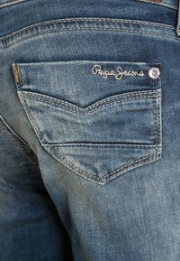 Los vaqueros de mezclilla azul cuentan con un bolsillo trasero de diseño en forma de V, superficie texturizada y "Pepe Jeans" bordado con hilo claro. Incluyen herrajes decorativos.