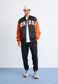 Man wearing black and orange Jordan jacket, black Jordan sweatpants, white sneakers, and a white cap standing against a plain background.