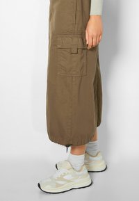 Khaki midi sukně s patkovou kapsou a stahovacími okraji, spárovaná se světle šedými ponožkami a pastelově žlutobílými teniskami s černou podrážkou.