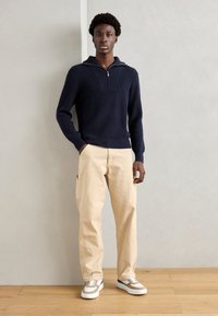 Σκούρο μπλε ριγέ πλεκτό πουλόβερ με μισό φερμουάρ, συνδυασμένο με μπεζ cargo παντελόνι και γκρι-λευκά αθλητικά παπούτσια, σε ξύλινο πάτωμα.