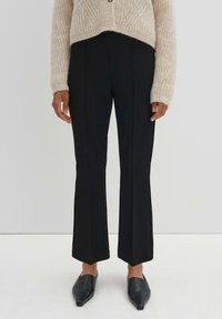 Pantalons noirs évasés avec des coutures frontales, fabriqués à partir d'un tissu lisse. Assorti à un pull beige léger et texturé et des chaussures noires à bout pointu.