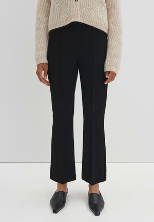 Black flared trousers with front seams, made from a smooth fabric. Paired with a light, textured beige sweater and pointed black shoes.